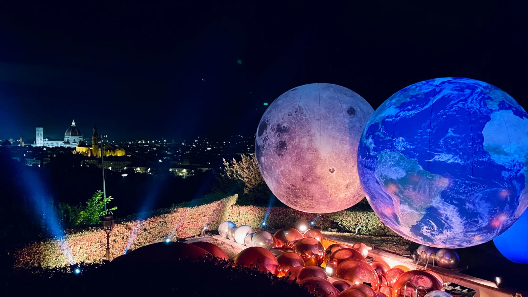 Panoramic evening view of Florence skyline with the Duomo and a wedding installation featuring large glowing Earth and Moon spheres in a villa pool.